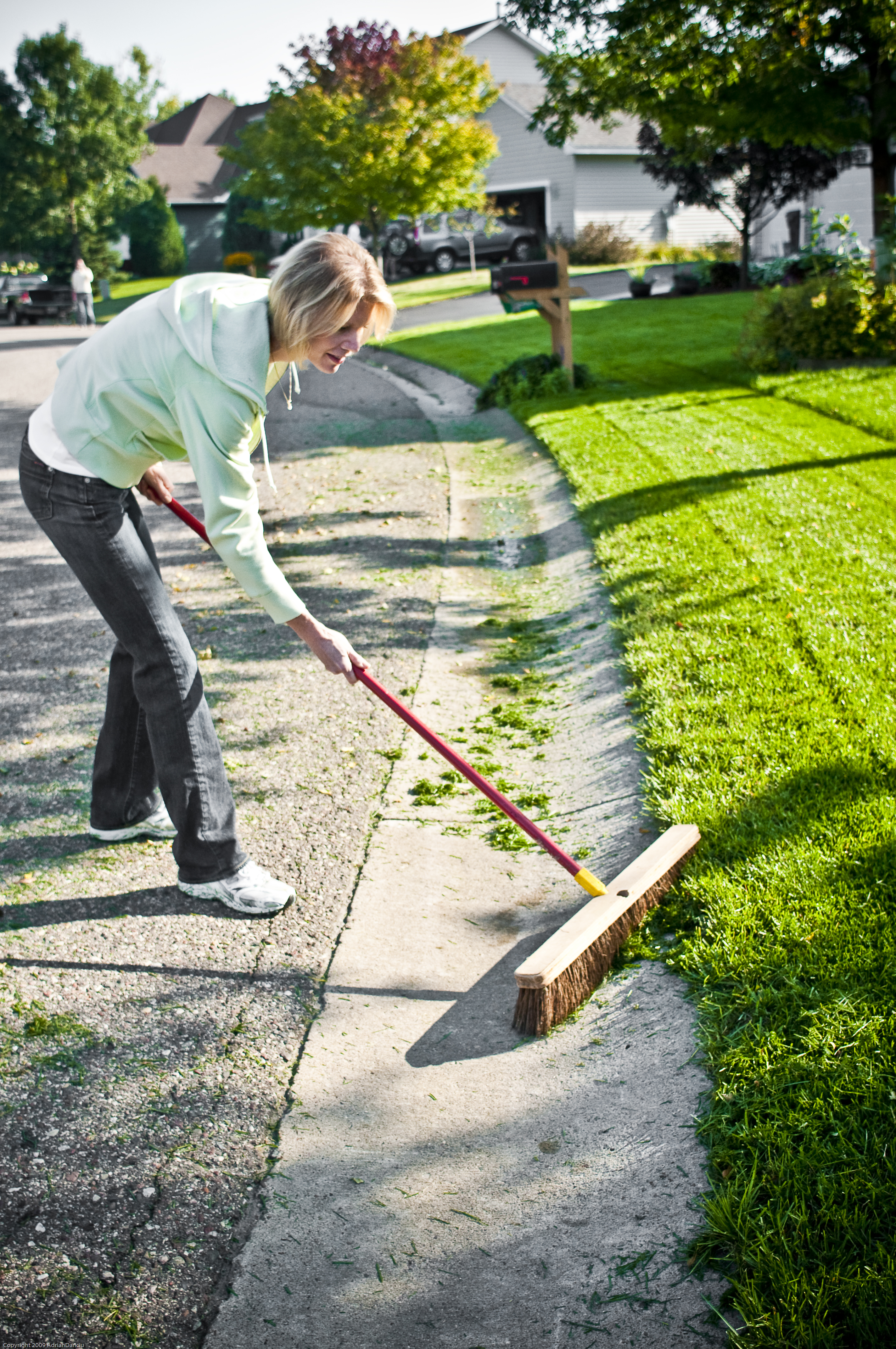 A middle-aged woman in a light green hoodie, blue jeans, and white sneakers sweeps lawn clippings from the side of the road with a wide broom.